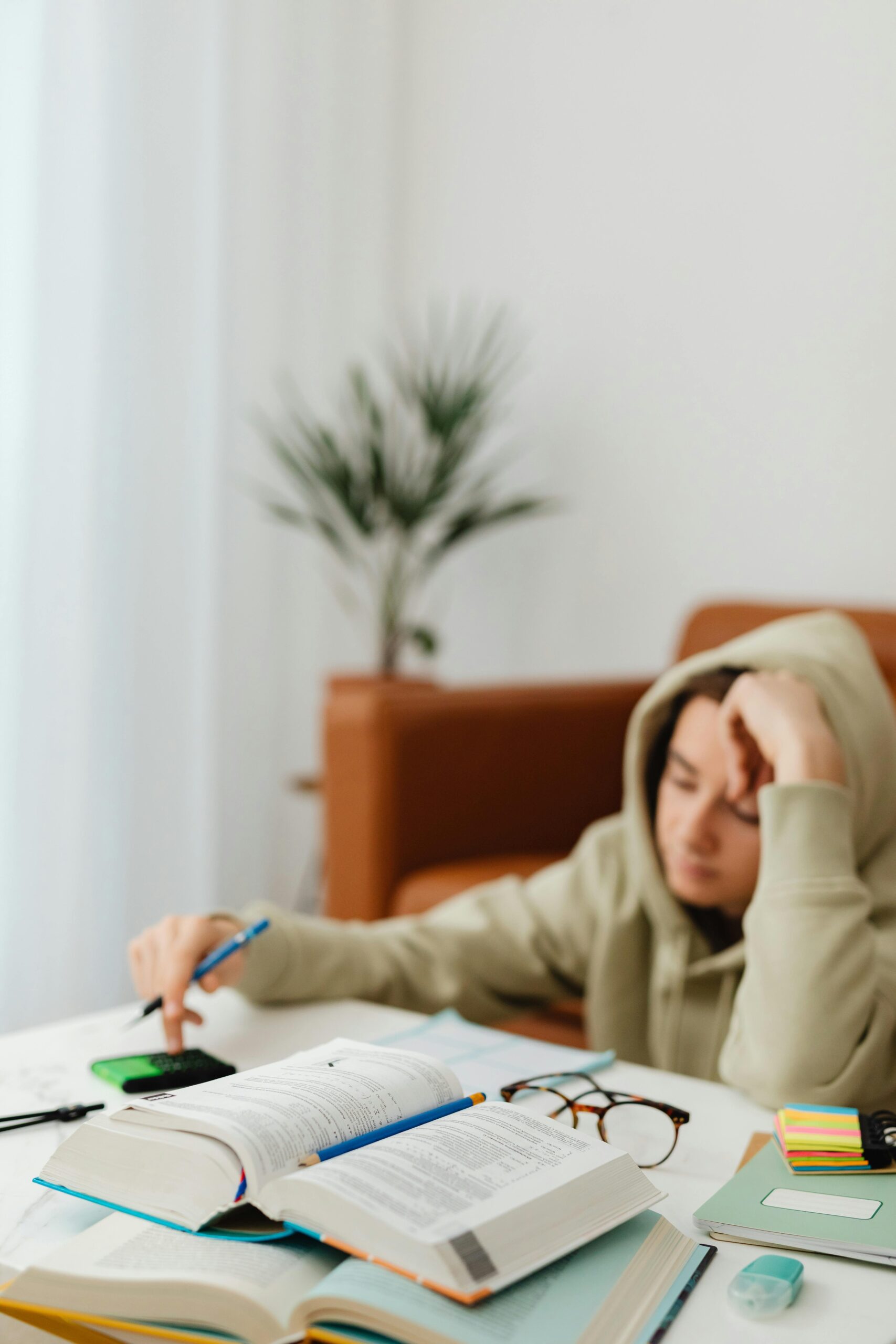 A teenager in a hoodie studying at home, surrounded by books and stationery.